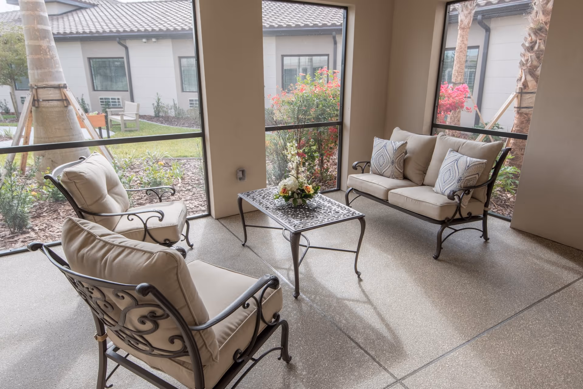 A screened-in seating area with cushioned metal chairs, a loveseat, and a decorative coffee table overlooking a landscaped courtyard.