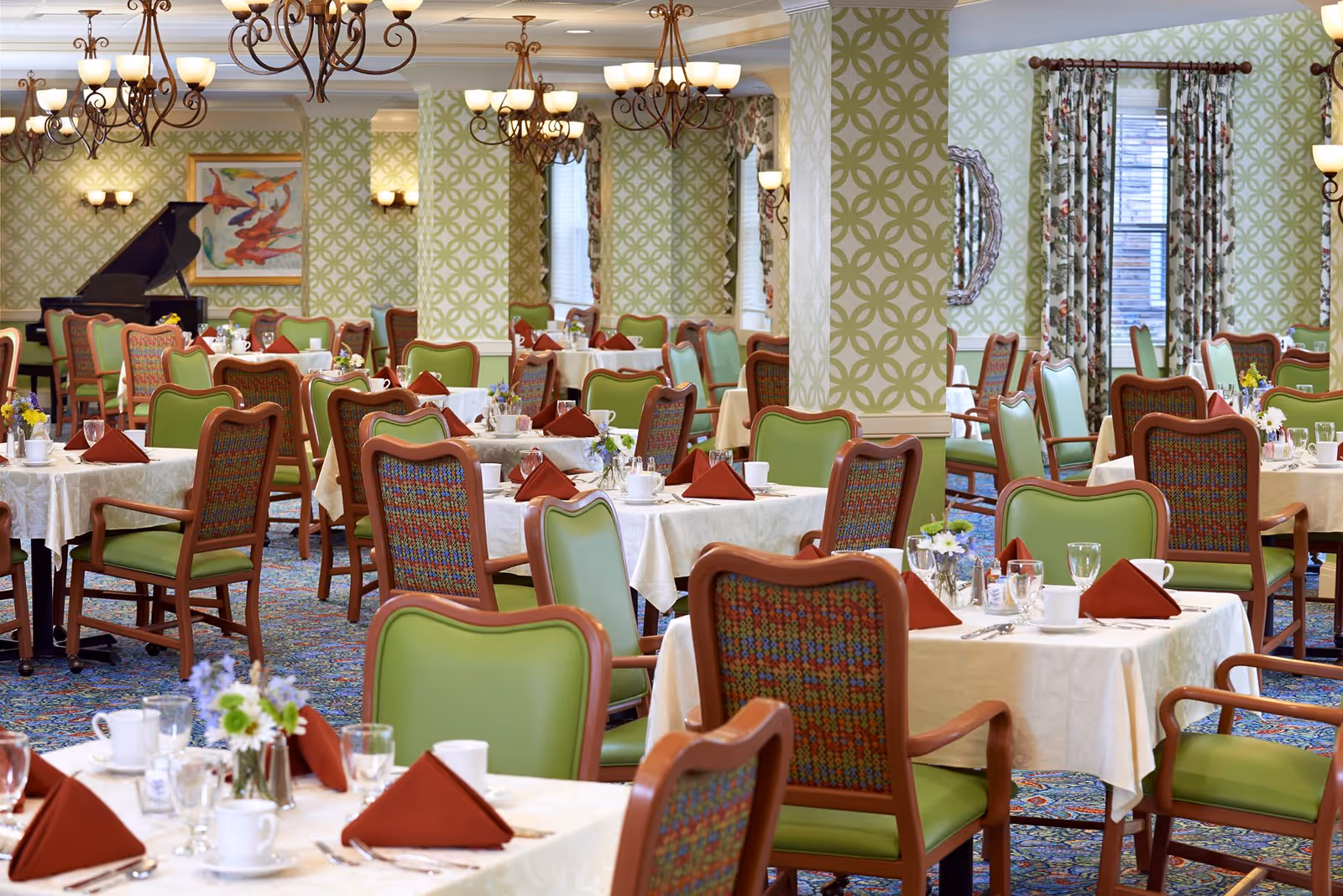 A spacious dining room with multiple tables covered in white tablecloths, each set with red folded napkins, cups, glasses, and silverware. The chairs have green cushions and wooden frames with patterned backs. The room features patterned green wallpaper, floral curtains, chandeliers, and a grand piano in the background.