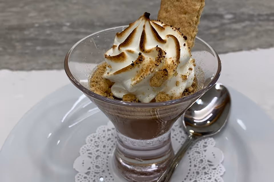 A small glass dessert cup of chocolate mousse topped with toasted whipped cream, graham cracker crumbs and a cookie, served on a white plate with a spoon.