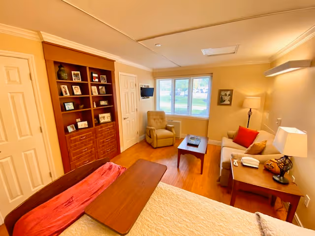 A cozy bedroom in a senior living facility featuring a bed with a wooden tray, a beige armchair near a window with blinds, a wooden coffee table, a beige sofa with red and beige cushions, a side table with a lamp and a phone, and a built-in wooden shelving unit with framed photos and decorative items.