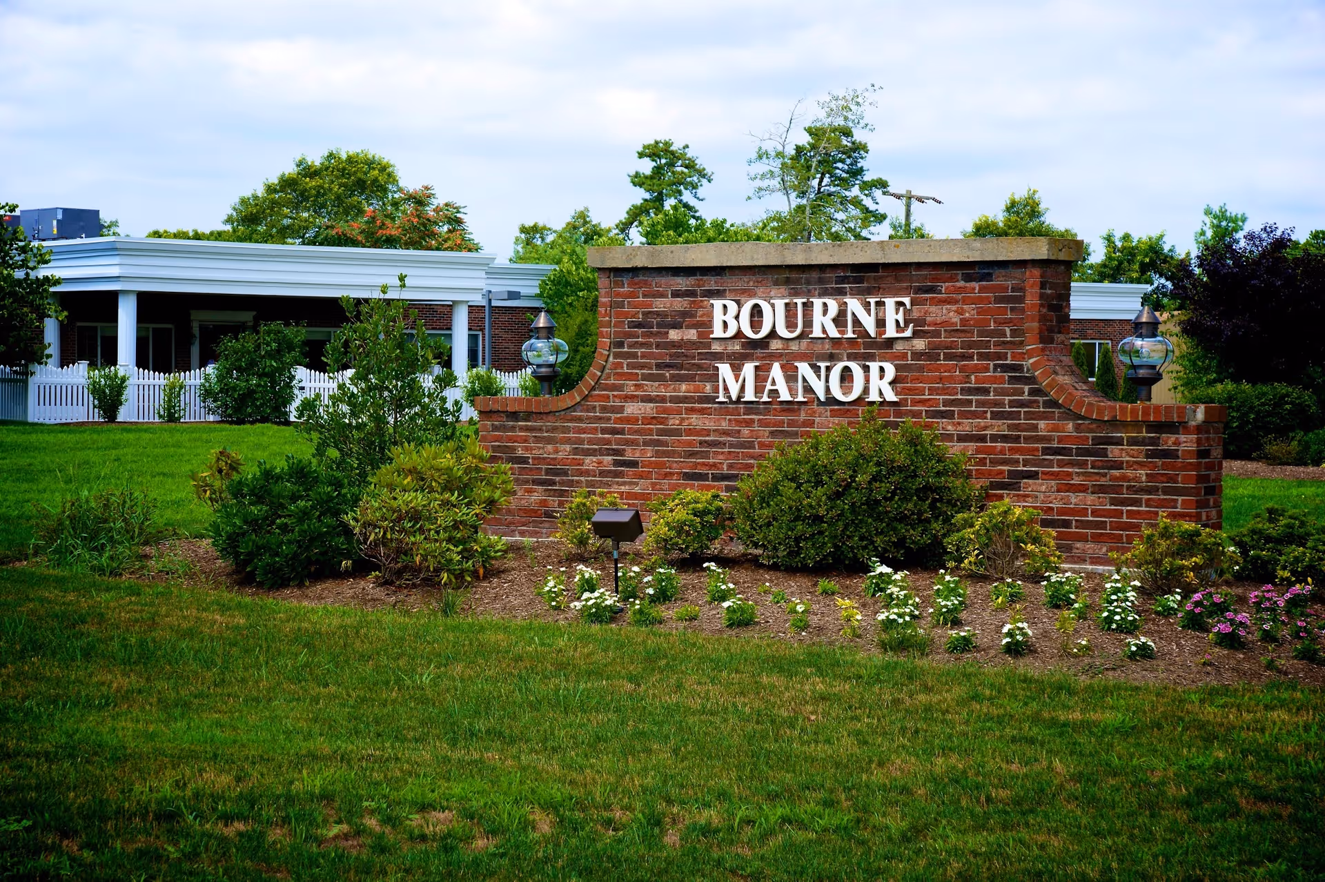 Brick entrance sign reading 'Bourne Manor' surrounded by landscaped shrubs and lawn with a low white-fenced building in the background.