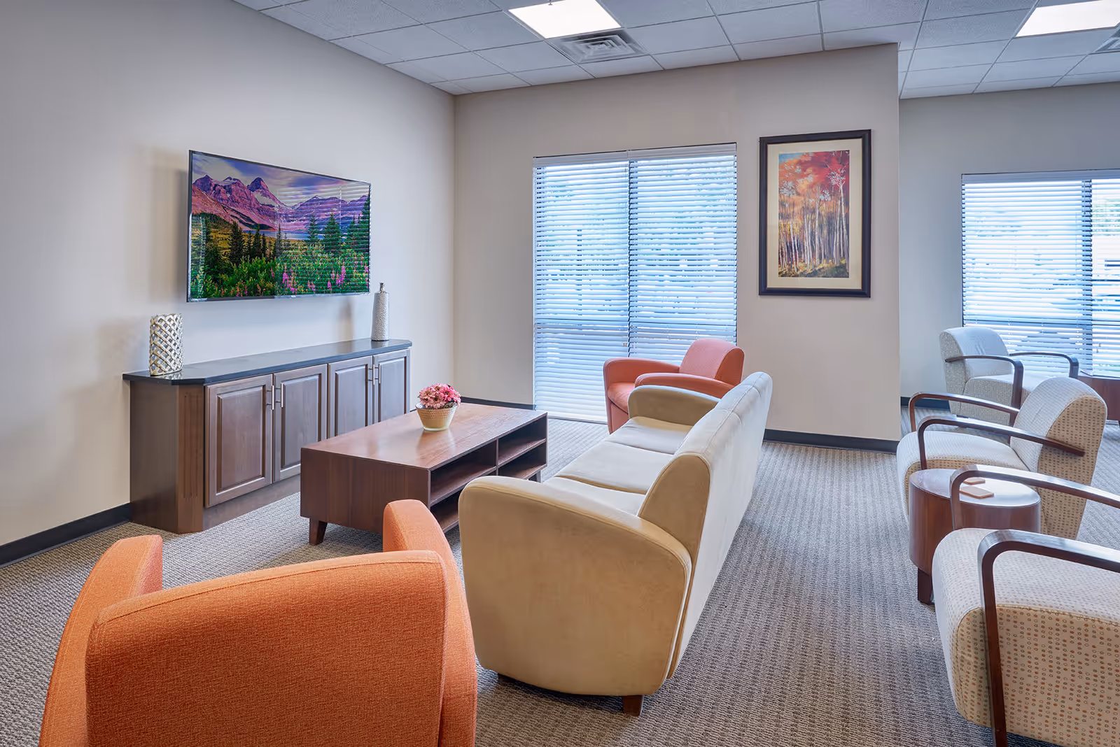 A well-lit living room with a beige sofa, two orange armchairs, and three patterned armchairs with wooden armrests. A wooden coffee table with a small flower pot is in the center. A flat-screen TV displaying a mountain landscape is mounted on the wall above a wooden cabinet. Two windows with blinds and a framed autumn forest painting are visible on the walls.