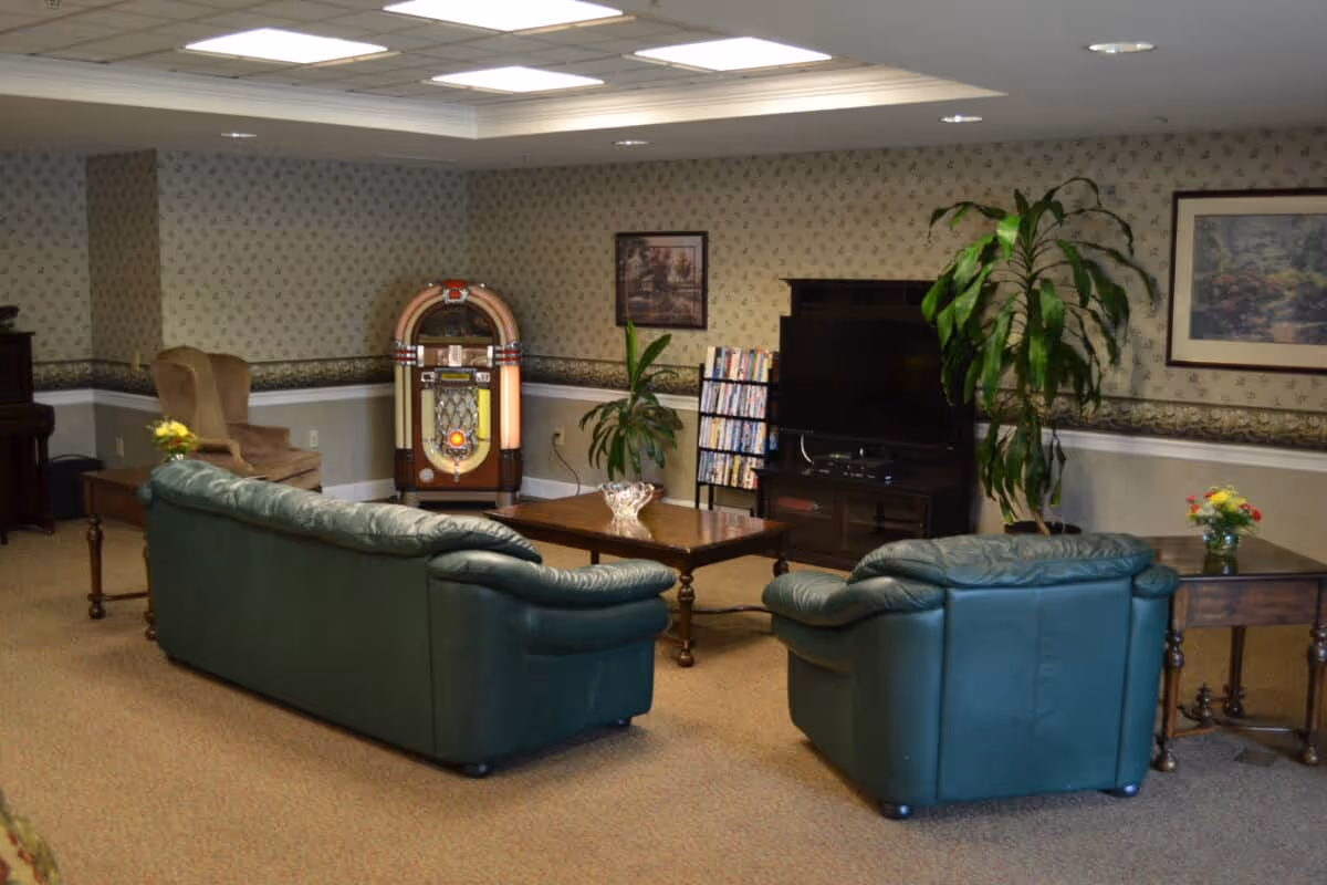 A cozy senior living common area with two green leather sofas and a matching armchair arranged around a wooden coffee table. There is a vintage jukebox against the wall, a TV on a stand with a DVD rack beside it, and two framed pictures hanging on the wallpapered walls. A large potted plant and small flower arrangements on side tables add a touch of greenery and color to the room.