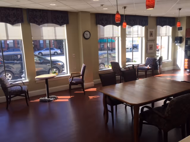 A sunlit room with large windows showing parked cars outside. Inside, there are several chairs arranged around small round tables near the windows and a larger wooden table with chairs in the center. The room has wooden flooring, beige walls, and red pendant lights hanging from the ceiling.