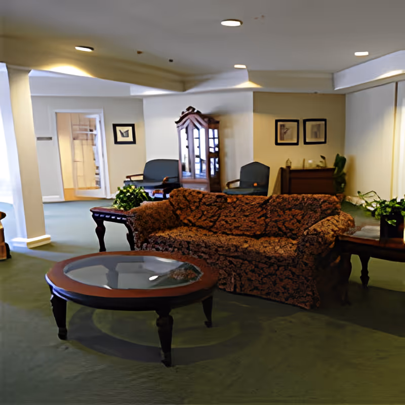 A cozy living room area in Heritage Assisted Living featuring a patterned sofa, a round glass-top coffee table, two armchairs, a wooden cabinet with glass doors, framed artwork on the walls, and several potted plants. The room has green carpet and soft overhead lighting.