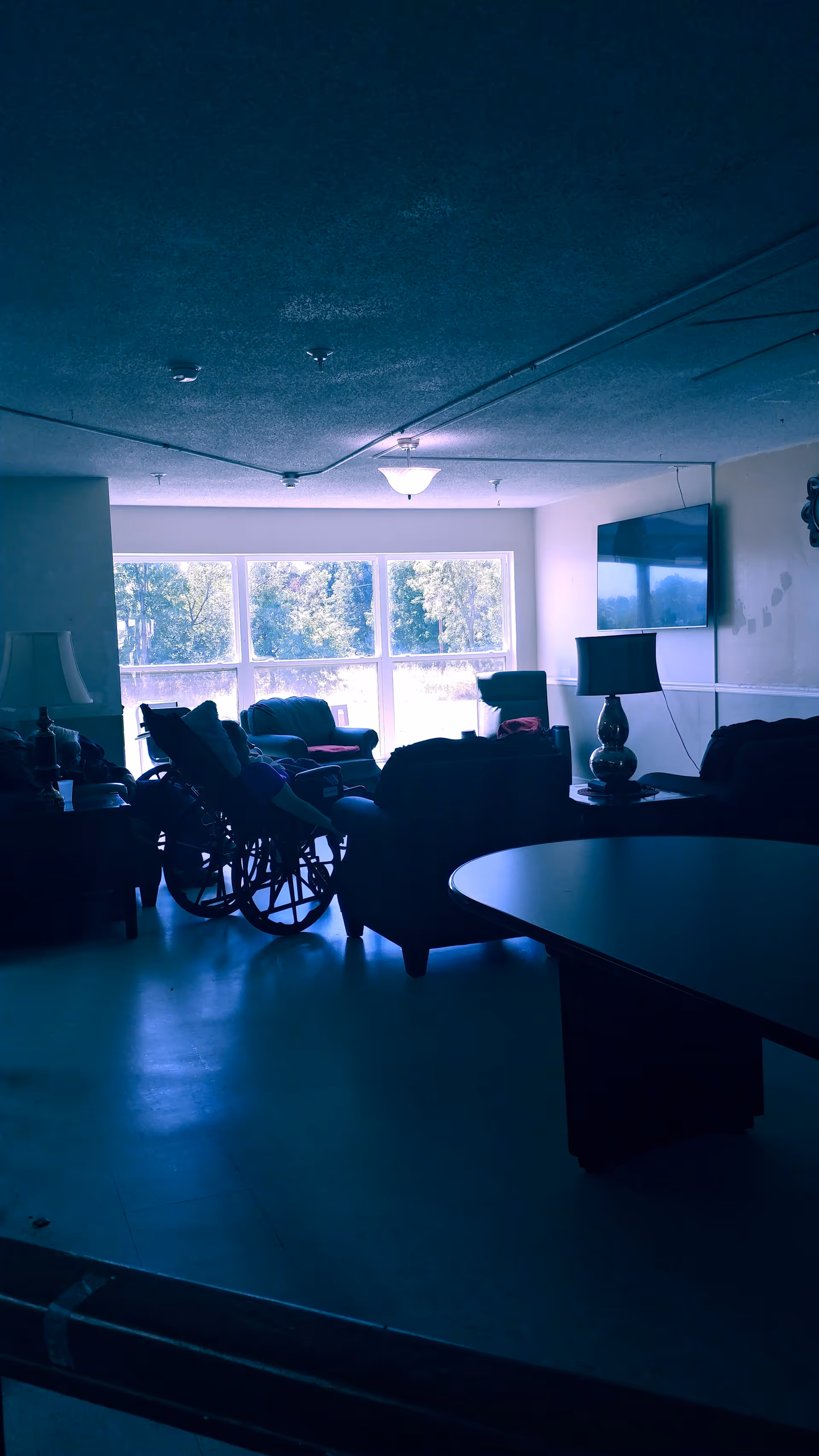 A dimly lit living room area with several armchairs, a wheelchair, a table with a lamp, and a wall-mounted television. Large windows in the background let in natural light and show trees outside.