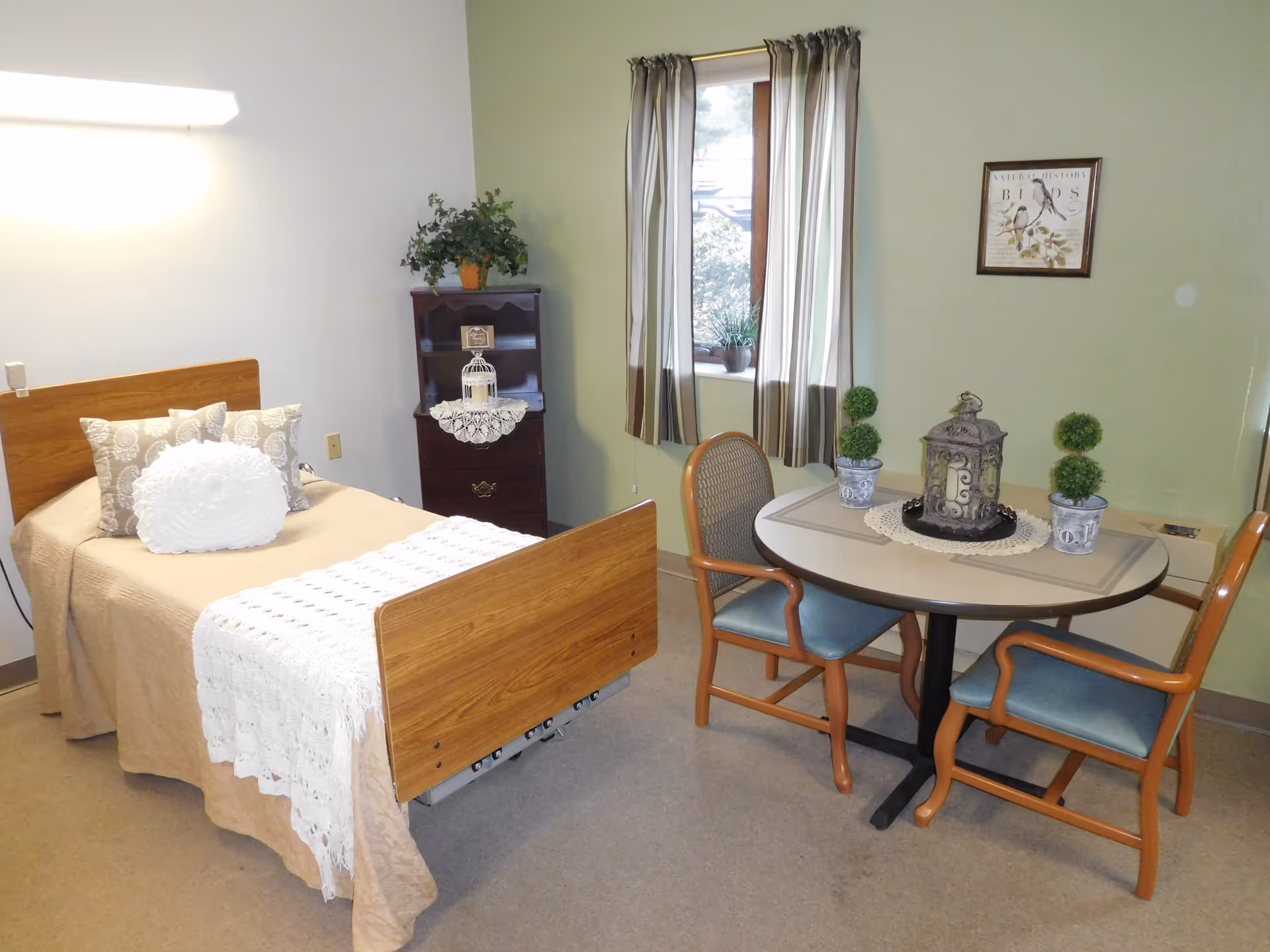 A cozy room with a single wooden bed covered with beige bedding and decorative pillows. Next to the bed is a small dark wooden cabinet with a plant and decorative items on top. A round table with two chairs is placed near a window with striped curtains. The table has decorative plants and a lantern centerpiece. The walls are painted light green and white, and a framed bird illustration hangs on the wall.