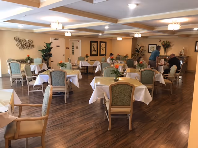 A dining room in a senior living facility with several tables covered in white tablecloths, each adorned with a small floral centerpiece. The room has wooden flooring, beige walls, and soft lighting from ceiling fixtures. There are upholstered chairs around the tables, and a few people are seated and interacting near the back of the room. Decorative plants and framed artwork are visible on the walls.
