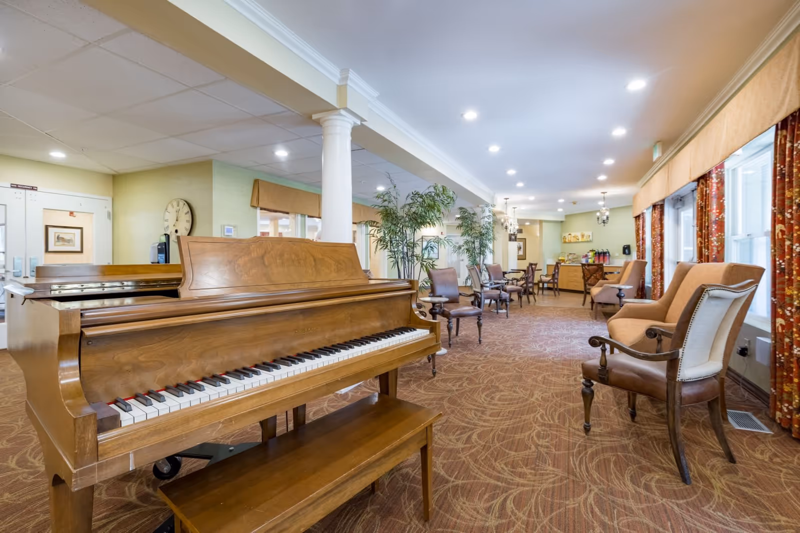 Spacious senior living common room with a wooden piano in the foreground and chairs and tables arranged along windows.