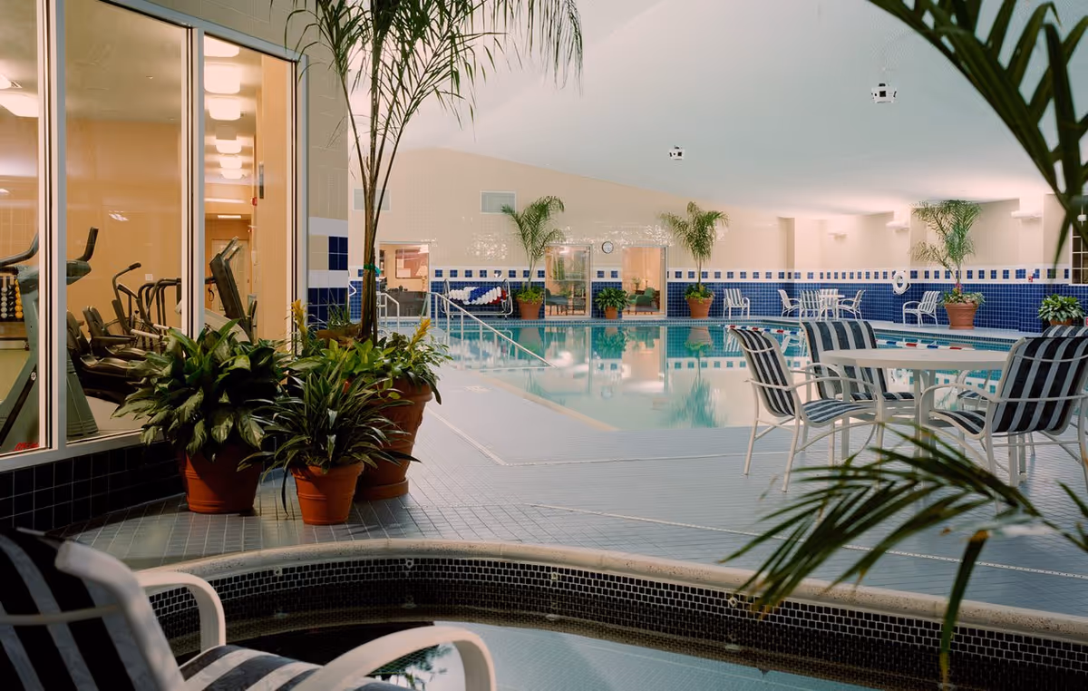Indoor swimming pool area with blue and white tiled walls, several potted plants, and striped chairs around tables. A fitness room with exercise equipment is visible through large glass windows on the left side.