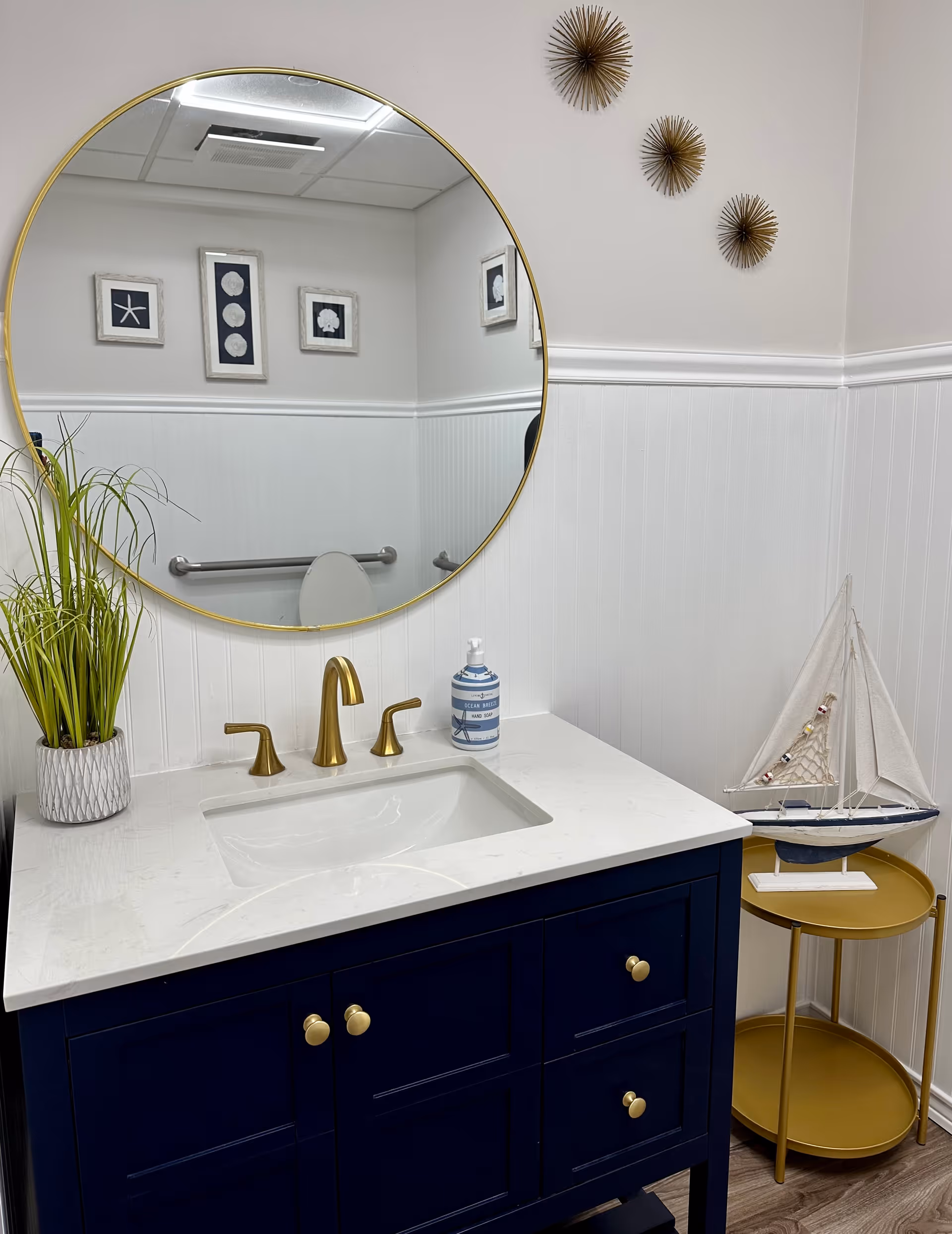 Modern bathroom vanity with a round mirror, navy cabinet, gold fixtures, decorative wall accents and a small sailboat on a side table.