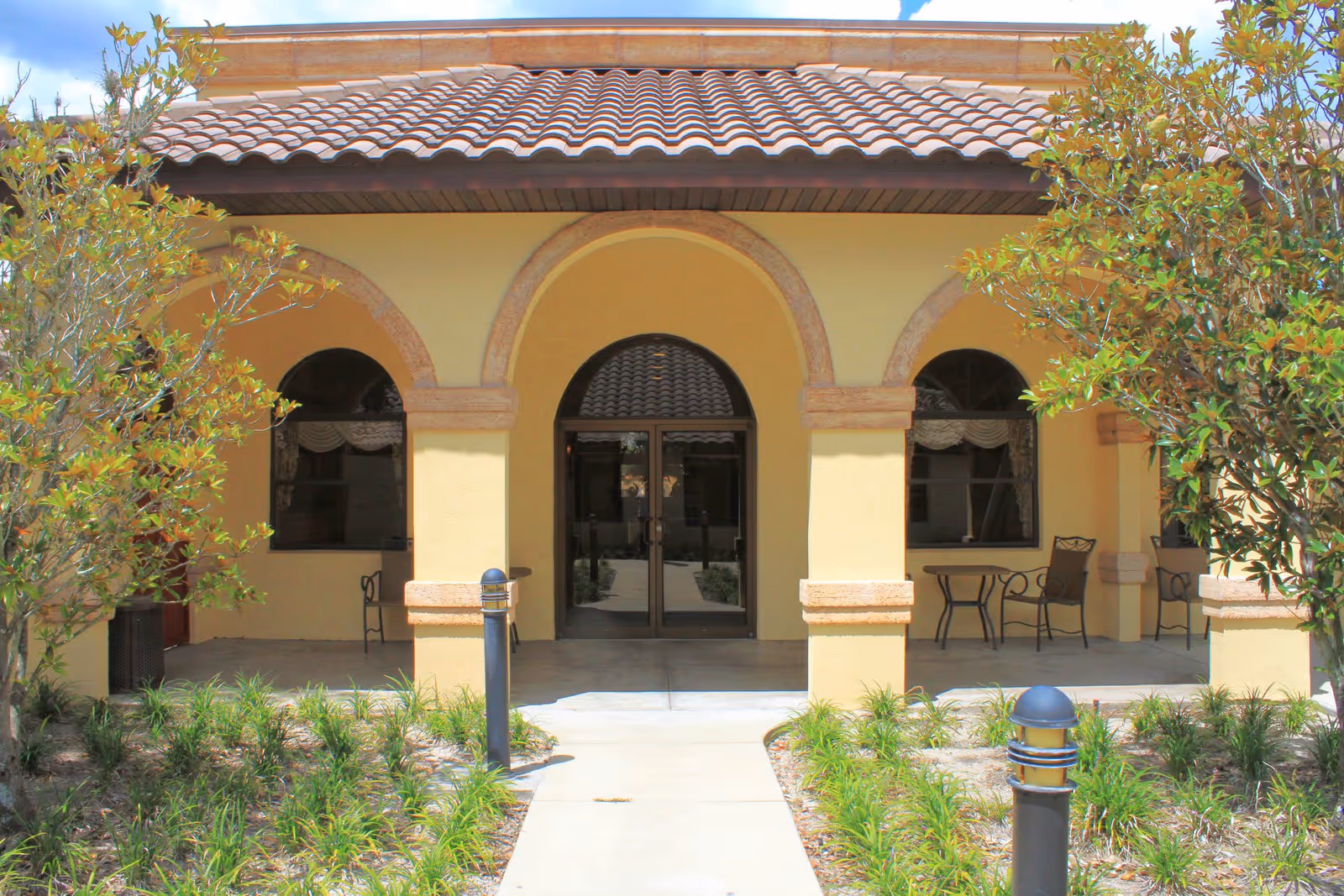 Front entrance of a building with a tiled roof and three arched openings. There are glass double doors in the center arch, flanked by windows with curtains. Two small tables with chairs are placed under the arches on either side. The walkway leading to the entrance is bordered by green plants and two black outdoor lights.