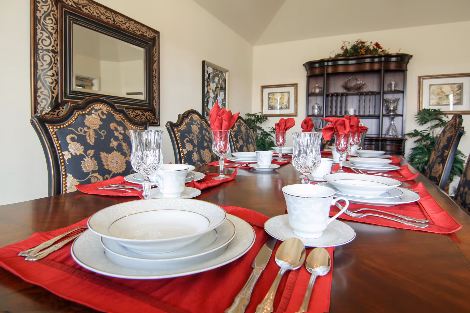 A formal dining room table set with white china plates, bowls, cups, silverware, and crystal glasses on red placemats and napkins. The table is surrounded by ornate chairs with floral upholstery. In the background, there is a wooden china cabinet with decorative items and framed artwork on the walls.