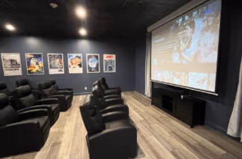 A small home theater room with two rows of black leather recliner chairs facing a large projector screen. The walls are dark blue and decorated with framed movie posters. The floor is light wood, and the room has a cozy, modern ambiance.