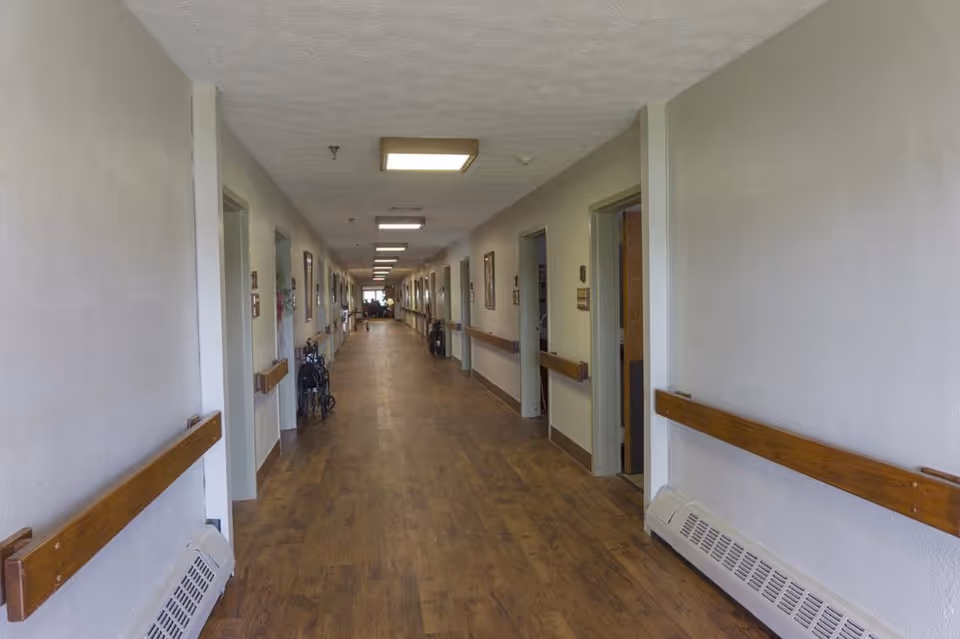 Long hallway in a senior living facility with wooden handrails on both sides, several open doorways, framed pictures on the walls, and a wooden floor. The hallway is well-lit with ceiling lights and has some mobility aids like walkers placed along the walls.