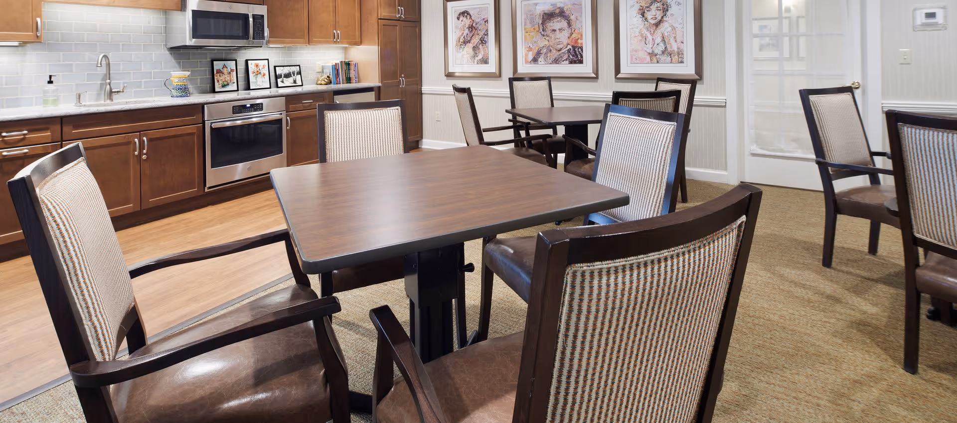 Communal dining area with several wooden tables and upholstered chairs adjacent to a small kitchen with stainless steel appliances and wood cabinets.