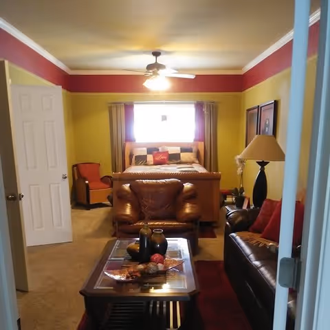 A cozy bedroom with a large bed against the back wall, flanked by curtains and a window. In front of the bed is a brown leather armchair and a glass-top coffee table with decorative items. To the right is a dark brown leather sofa with red cushions and a tall black lamp with a beige lampshade. The walls are painted yellow with a red border near the ceiling, and the room is softly lit by a ceiling fan light.