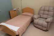 Small resident room with a single bed, bedside cabinet and IV pole, and a padded recliner chair.