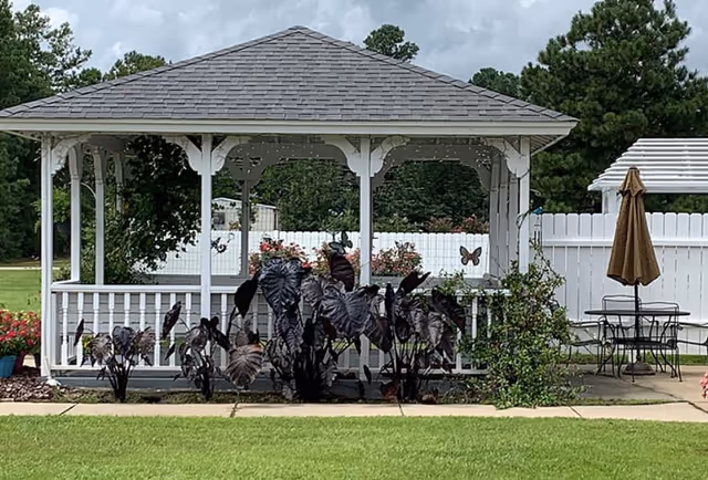 A white wooden gazebo with a shingled roof situated in a garden area with green grass and various plants. There is a white fence in the background, and to the right of the gazebo, there is a patio table with an umbrella. Trees and shrubs are visible beyond the fence.