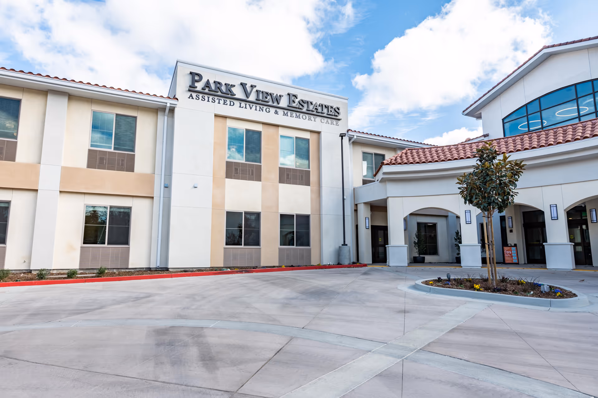 Exterior view of Park View Estates Assisted Living & Memory Care building with a clear sky and a small landscaped area with a tree in front of the entrance.