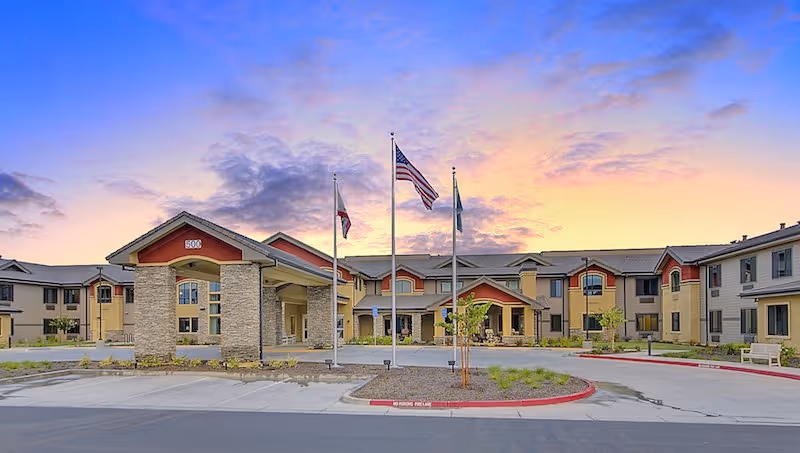 Front exterior of a two-story senior living community with a covered porte-cochère, three flagpoles, and a colorful sunset sky.
