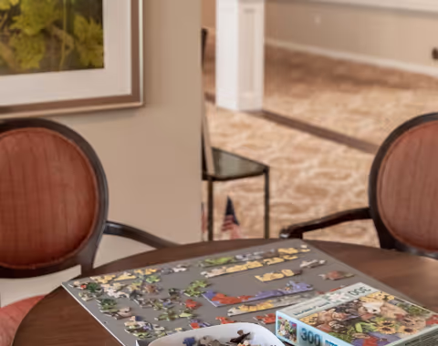 A round wooden table with a partially completed jigsaw puzzle and a puzzle box on it, surrounded by upholstered chairs with reddish cushions. The background shows a carpeted floor and a wall with a framed picture.