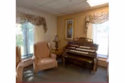 Small common room with upholstered armchairs, an organ, a side table with a lamp, and windows with valanced curtains.