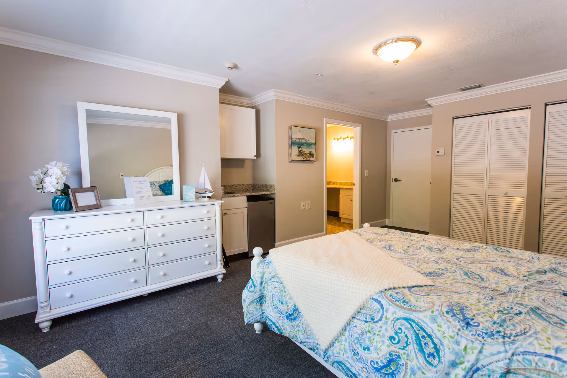 A bright and clean bedroom in Magnolia Manor Assisted Living featuring a bed with a colorful paisley-patterned bedspread and a cream-colored throw blanket. There is a white dresser with a large mirror on top, decorated with a vase of white flowers, a small framed picture, and a decorative sailboat. The room has beige walls, a gray carpet, and a ceiling light fixture. A doorway leads to a bathroom with a granite countertop and illuminated vanity mirror. There are white louvered closet doors and a closed door on the far wall.