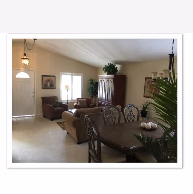 A cozy living and dining area with beige walls and carpet. The living room features a brown sofa, two armchairs, a wooden cabinet with a plant on top, and a floor lamp near a window with blinds. The dining area has a dark wooden table with four chairs and a decorative centerpiece. There are framed pictures on the walls and a hanging light fixture above the dining table.