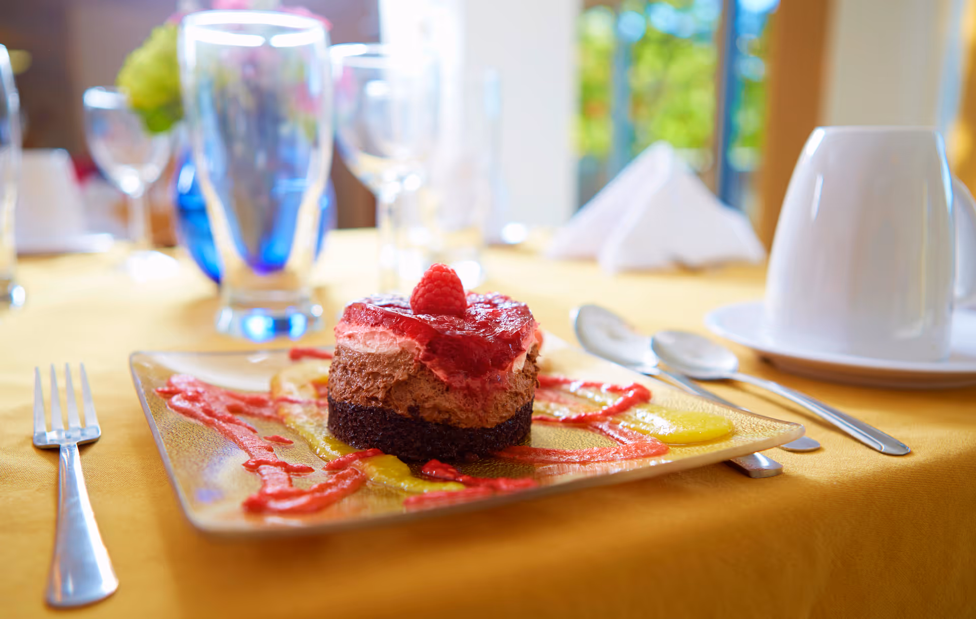 A close-up of a plated dessert with layers of chocolate and raspberry mousse topped with a raspberry, served on a square glass plate with red and yellow sauce drizzled around it. The table is covered with a yellow tablecloth and set with a fork, spoons, a white cup on a saucer, and several glasses in the background.