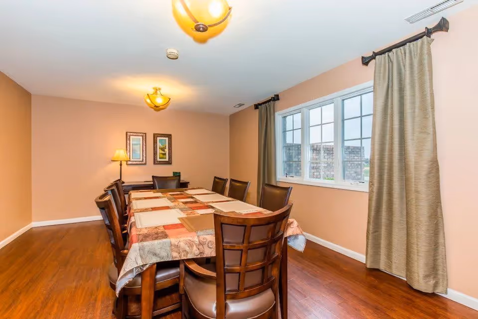 A dining room with a rectangular wooden table covered with a patchwork tablecloth and surrounded by eight wooden chairs with cushioned seats. The room has light brown walls, a wooden floor, two framed pictures on the far wall, a table lamp, and a window with beige curtains on the right side.