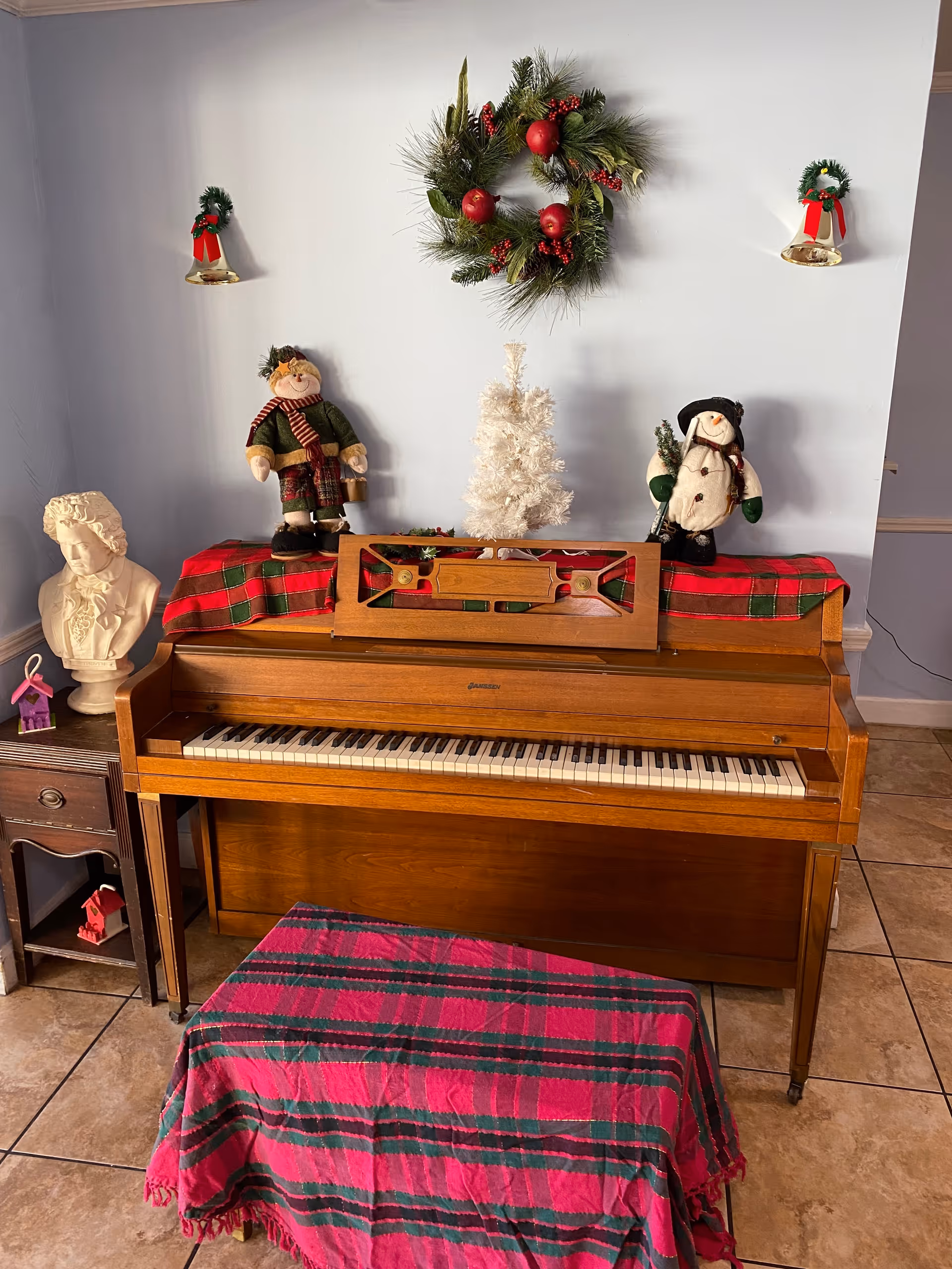 A wooden upright piano decorated for Christmas with a red and green plaid cloth on top and a matching cloth covering the bench. On top of the piano are a small white Christmas tree, a snowman doll, and a doll dressed in winter clothes. Above the piano hangs a Christmas wreath with red ornaments, and two small wreaths with red ribbons are mounted on the wall on either side. To the left of the piano is a small wooden side table with a white bust sculpture and a small purple house decoration.