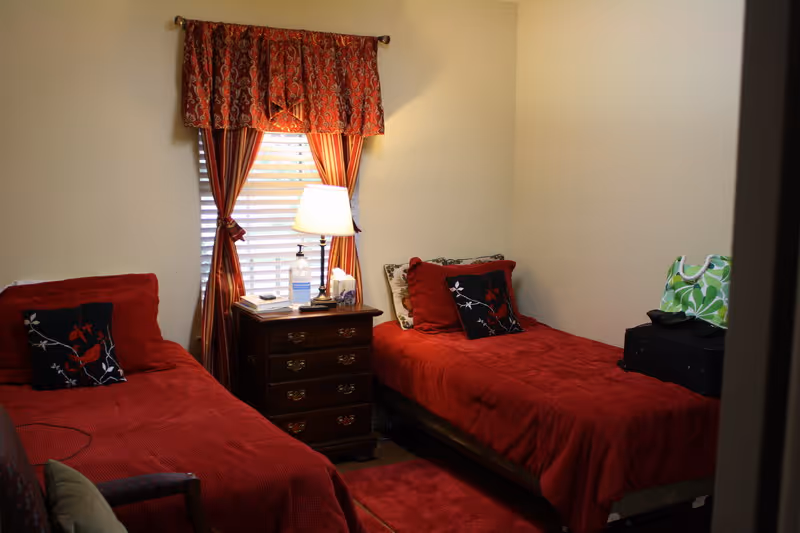 A small bedroom with two single beds covered in red bedspreads and decorative pillows. Between the beds is a wooden nightstand with a lamp, a bottle of hand sanitizer, and some tissues. A window with red patterned curtains is behind the nightstand. There is a green and white patterned bag on a black suitcase on the bed to the right.