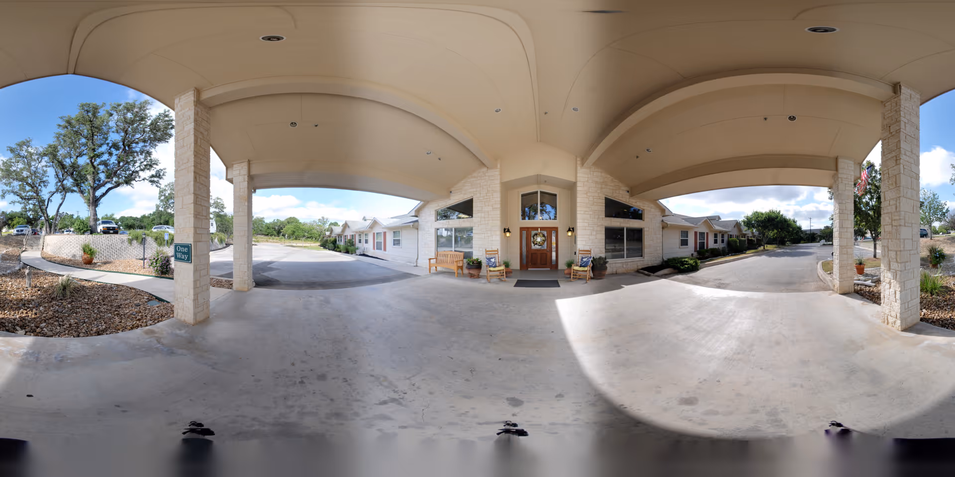 Covered entrance area of a senior living facility with stone pillars and a concrete driveway. There are benches and potted plants near the entrance door. The surrounding area includes landscaped sections with rocks and trees under a partly cloudy sky.