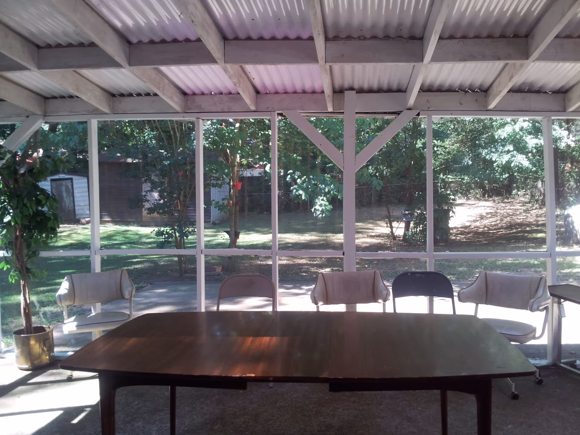 A covered outdoor patio area with a wooden table and five chairs arranged around it. The patio is enclosed with a screen, and outside there is a grassy yard with trees and a small shed visible in the background.