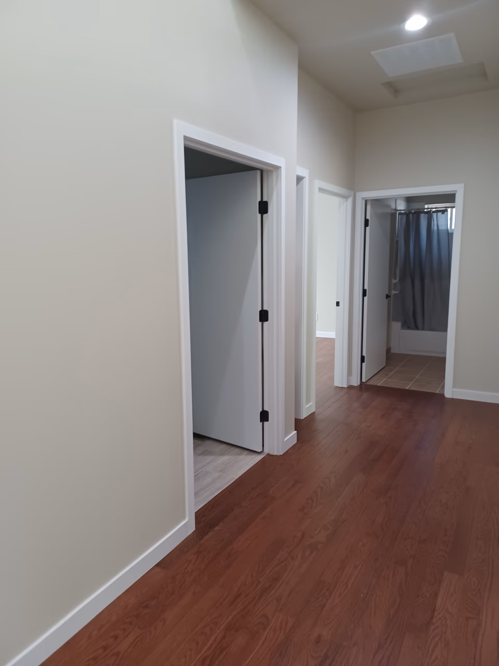 Hallway with wood flooring and several open doorways, one leading to a bathroom with a shower curtain.