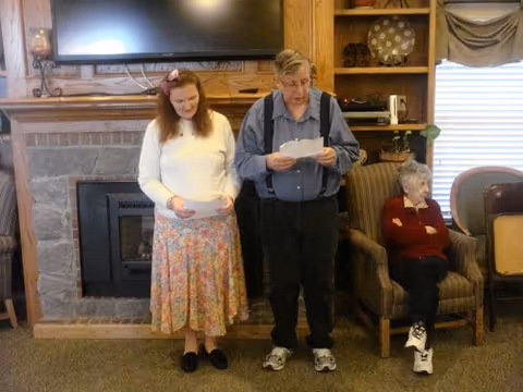 Two people standing and reading and one person seated in a common room in front of a fireplace and TV.