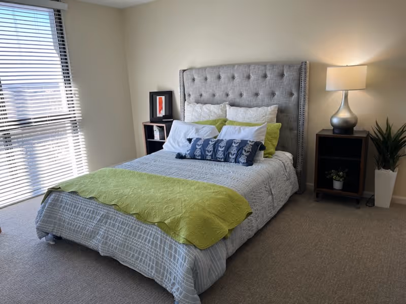 A neatly made bed with a tufted gray headboard in a bedroom. The bed has white and green pillows, a white and gray bedspread, and a green throw blanket at the foot. There are two dark wooden nightstands on either side of the bed, one with a silver lamp and the other with a framed picture. A potted plant is placed on the floor next to the right nightstand. A window with blinds is on the left side of the room, letting in natural light.