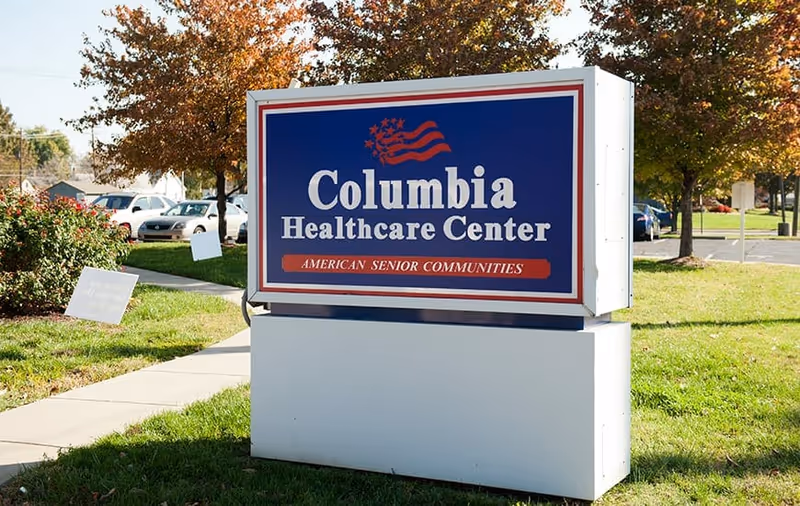 Outdoor sign for Columbia Healthcare Center with the tagline American Senior Communities, surrounded by grass, trees with autumn leaves, and parked cars in the background.