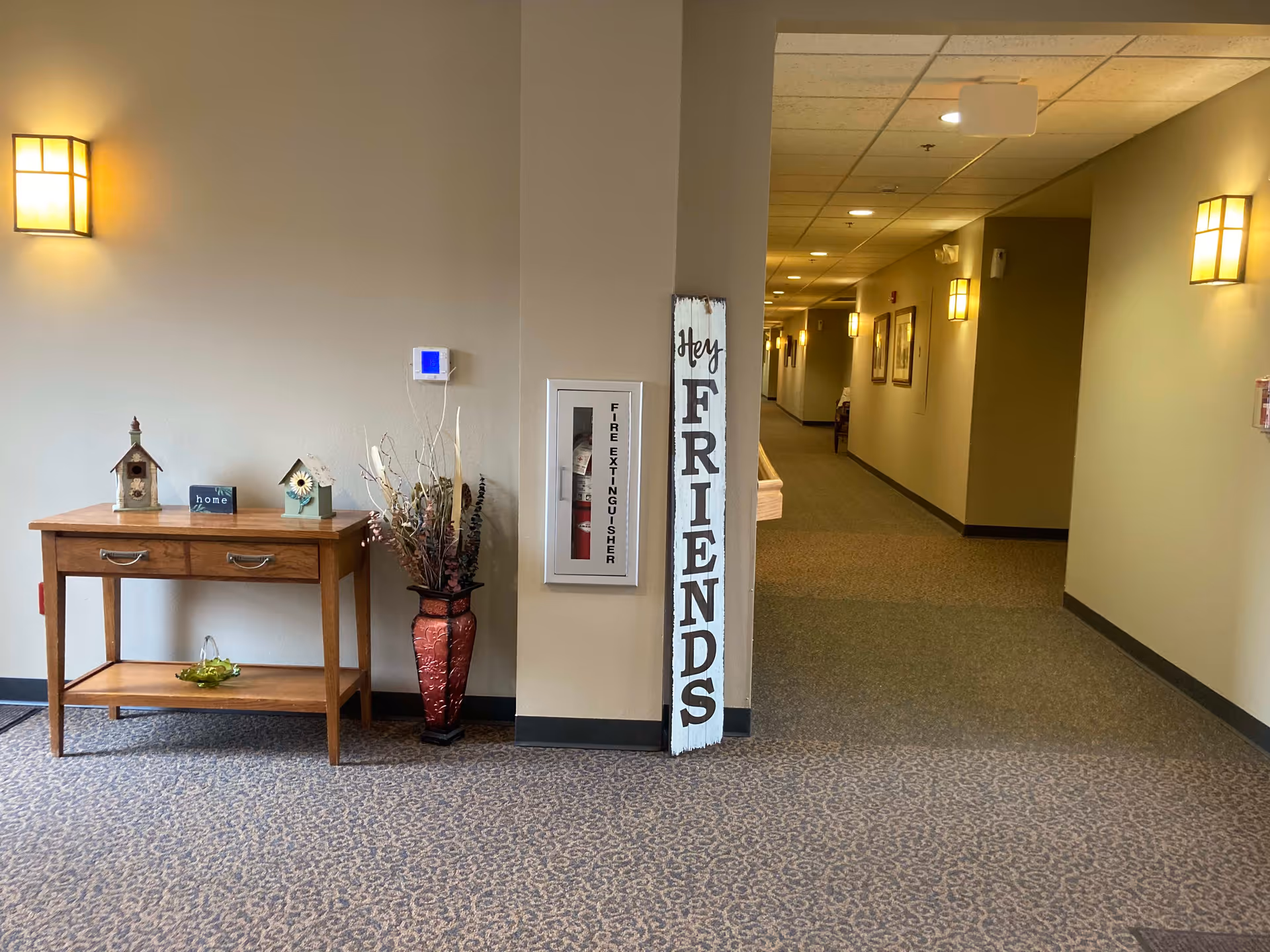 Interior hallway of a senior living facility with beige walls and carpeted floor. On the left side, there is a wooden table with decorative birdhouses and a small sign that says 'home.' Next to the table is a tall red vase with dried plants. A fire extinguisher is mounted on the wall. A vertical wooden sign leaning against the wall reads 'Hey FRIENDS.' The hallway is lit by wall sconces and ceiling lights, with framed pictures hanging along the corridor.