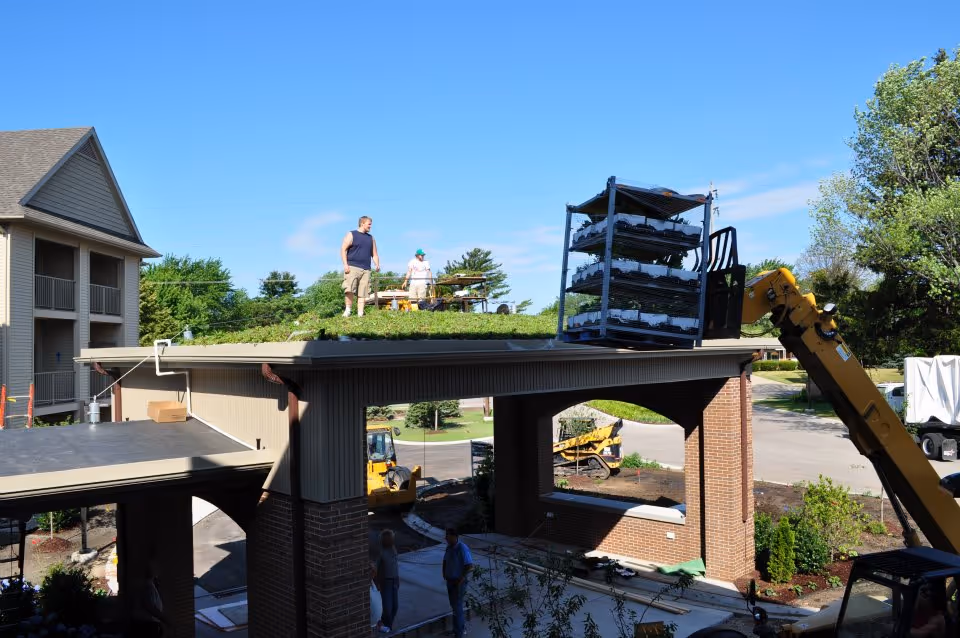 Workers installing a green roof on the covered entrance canopy of a brick senior living building with a crane and plant racks.