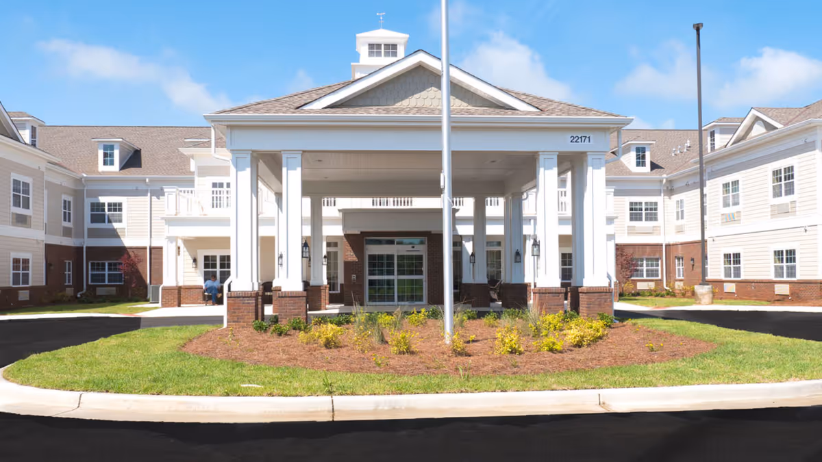 Front exterior view of The Goldton at Athens senior living facility with a covered entrance supported by white columns, landscaped greenery in front, and a clear blue sky above.
