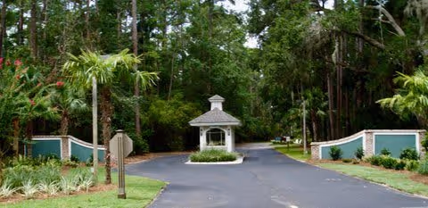 Entrance driveway to a facility with a small white guardhouse in the center, surrounded by greenery including palm trees and other plants, with a paved road leading into a wooded area.