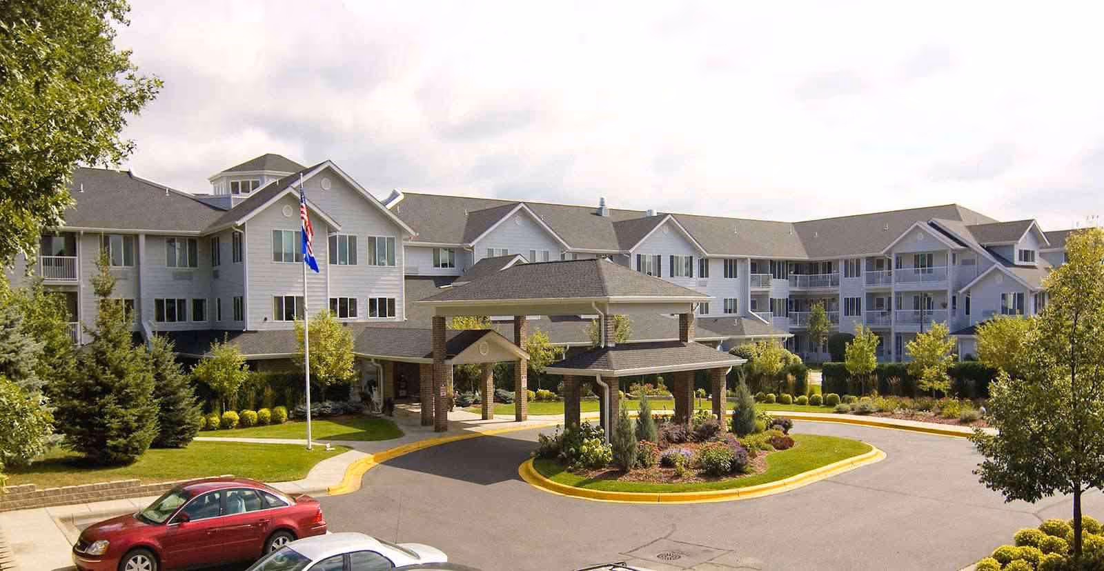 Exterior view of The Lodge at White Bear, a multi-story senior living facility with a covered entrance driveway, surrounded by landscaped greenery and trees, with a few parked cars in front.