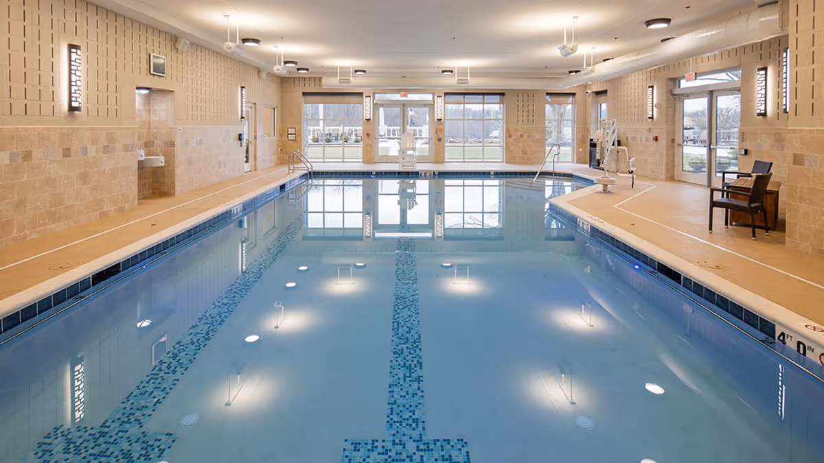 Indoor swimming pool with clear blue water, beige tiled walls, and large windows letting in natural light. There are poolside chairs, a pool lift for accessibility, and wall-mounted lights along the perimeter.