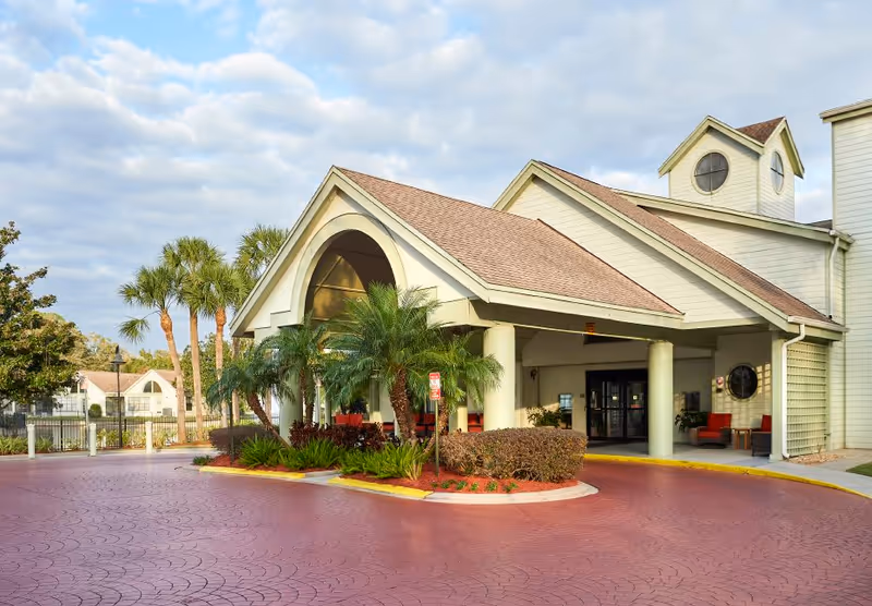 Front exterior view of a senior living facility named Cascade Heights with a covered entrance, palm trees, and a circular driveway under a partly cloudy sky.