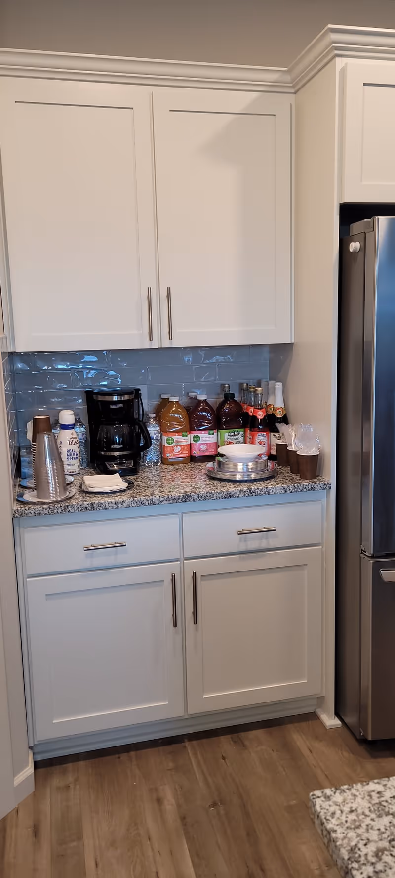 A kitchen counter with white cabinets above and below, a granite countertop holding a coffee maker, several bottles of juice, disposable cups, napkins, and utensils. A stainless steel refrigerator is visible on the right side.