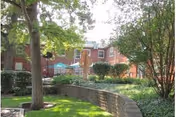Outdoor garden area with green grass, trees, bushes, and a curved brick retaining wall. In the background, there is a red brick building with windows and a patio area with blue umbrellas.