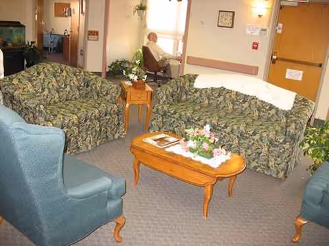 A cozy senior living lounge with floral sofas, coffee table, plants, and a resident seated.