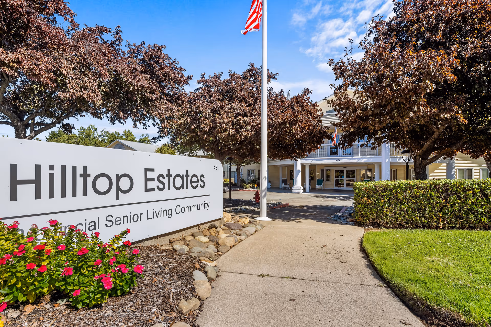 Entrance to Hilltop Estates, a provincial senior living community, featuring a large white sign with the community name, a paved walkway, manicured bushes, blooming flowers, trees with reddish leaves, an American flag on a flagpole, and the front exterior of the building in the background under a blue sky.